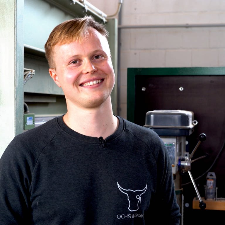 Ein junger, blonder Mann steht in einer Werkstatt und lächelt in die Kamera