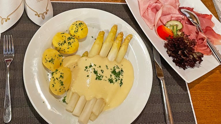 Weißer Spargel mit Sauce Hollandaise und Kartoffeln auf weißem Teller, dazu Schinken auf einer extra Platte