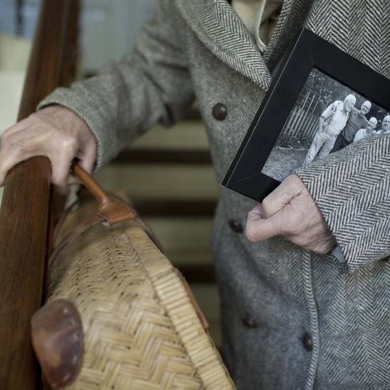 Seniorin mit Koffer und Familienfoto unter dem Arm steigt eine Treppe hinunter: Orientierungsstörungen gehören auf mit zur Demenz. Die sind am Anfang zwar nicht so deutlich, nehmen aber zu.