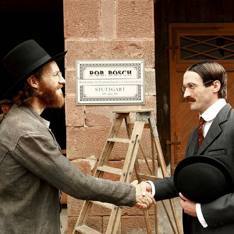 Zwei dunkel gekleidete Männer geben sich die Hand. Beide tragen Hut und Brille. Hinter ihnen sieht man mittig an einer Mauer ein Schild auf dem steht: "Rob Bosch, Werkstätte für Feinmechanik & Elektrotechnik, Stuttgart."