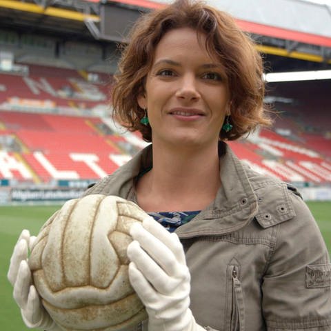 Lena Ganschow steht in einem leeren Stadion. Sie trägt Handschuhe und hält einen alten Fußball in den Händen.
