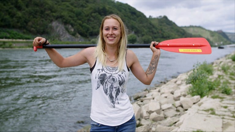 Sportliche, junge Frau steht mit Paddel vor dem Mittelrheintal