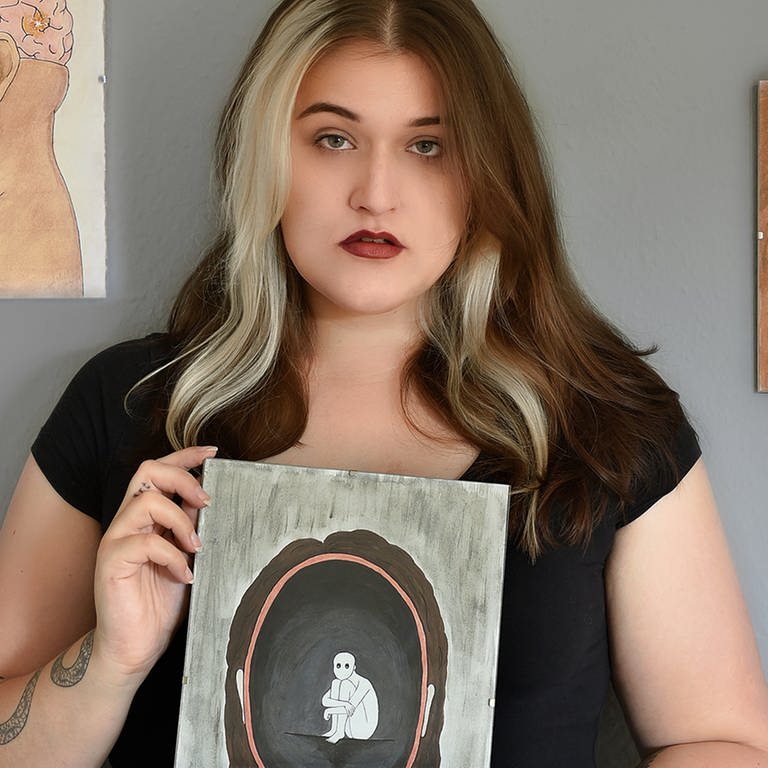 Junge Frau mit einem von ihr gemalten Bild in der Hand, vor einer Wand mit weiteren von ihr gemalten Bildern