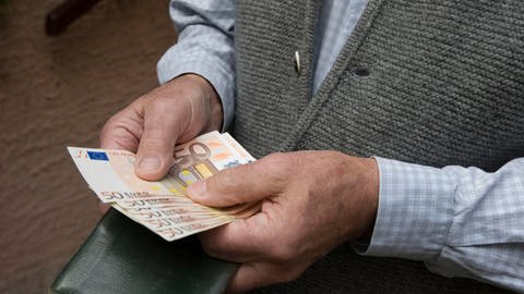 Ein älterer Mann mit Strickweste hält mehrere 50-Euro-Scheine aufgefächert in der Hand