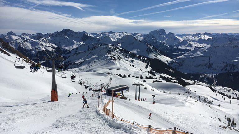 Skifahrer fahren im Skigebiet Damüls den Berg hinunter. Im Hintergrund sind schöne beschneite, weiße Berggipfel zu sehen.
