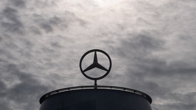 Ein Mercedes-Stern ist auf der Mercedes-Benz Hauptniederlassung in Stuttgart vor dunklen Wolken und Abluft, die aus einem Industrieschornstein kommt, zu sehen.