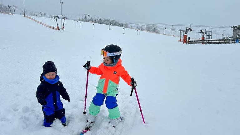 Zwei Kinder im Schnee, eines auf Skiern, im Hintergrund ein Skilift