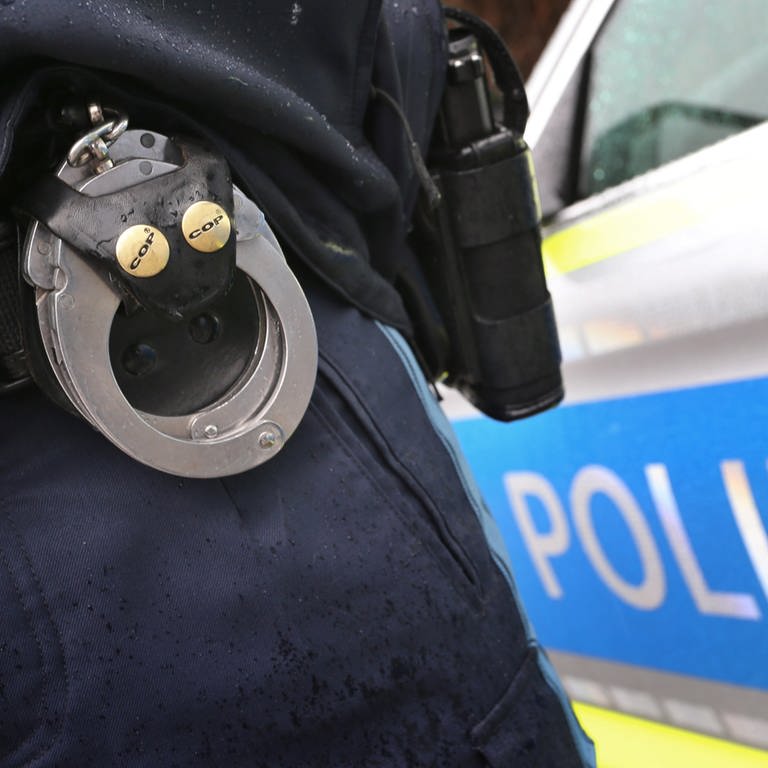 Ein Polizist mit Handschellen und einer Pistole am Gürtel steht vor einem Streifenwagen