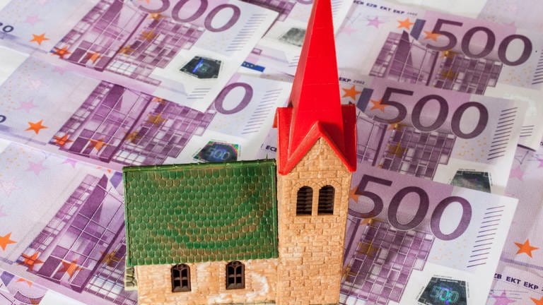 Eine Spielzeugkirche steht auf einem Untergrund auf 500-Euro-Scheinen. Geld wird im Bistum Trier knapp. Es muss Millionen Euro einsparen.