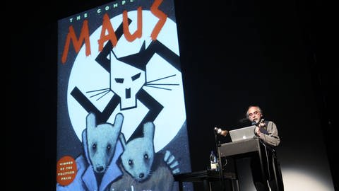 Bild eines älteren Mannes an einem Rednerpult, neben ihm projiziert das Buchcover von 'Maus'. Es zeigt einen Art stilisierten Hitler als Maus in einem Hakenkreuz.