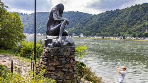 Bronze-Skulptur der Loreley auf einem Felsen bei Sankt Goar am Rhein. Ein Tourist fotografiert das Denkmal.