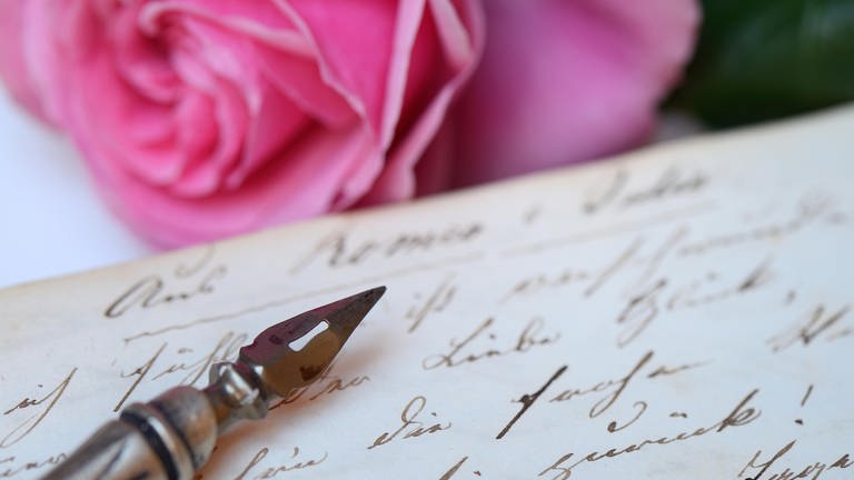 Federhalter mit Schreibfeder auf Buch mit alter Handschrift: Liebesbriefe sind Zeugnisse ihrer Zeit und Gesellschaft. Hieß es früher: „Ich liebe Dich“, werden heute Emojis mit Herzchen und Küsschen gesendet.
