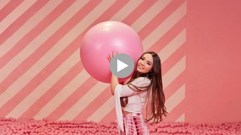 Die Teenager-Influencerin Jule Nagel aus Stuttgart ist in einem Bällchen-Bad mit rosa Bällen vor einem rosa-weiß-gestreiften Hintergrund. Sie ist bekleidet mit einer weißen bauchfreien Bluse und einer rosa-weißen Hose. Jule hält einen großen rosafarbenen Ball in beiden Händen über ihren Kopf