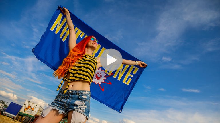 Eine Besucherin mit langen roten Haaren und kurzem gelb-schwaz geringelten Top und Shorts auf dem Festivalgelände mit sommerlichem blauen Himmel. Sie hält hinter sich eine blaue Fahne mit gelbem Aufdruck "Nature One"