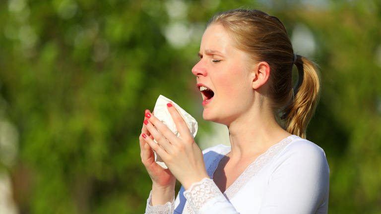 Eine junge Frau niest in der grünen Natur in ein Taschentuch: Niesen ist physiologisch ein explosives Atmen; die gleichen Körperpartien sind beteiligt wie beim normalen Atmen. Das "Nieszentrum" im Gehirn führt das Kommando.